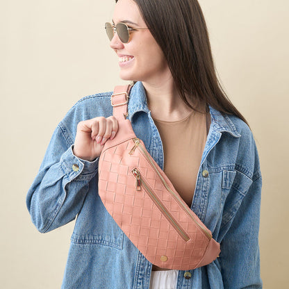 A young woman with long dark hair, sunglasses, and a denim jacket smiles while holding The Darling Effect’s Rose Trendy Luxe Belt Bag in woven pink vegan leather.