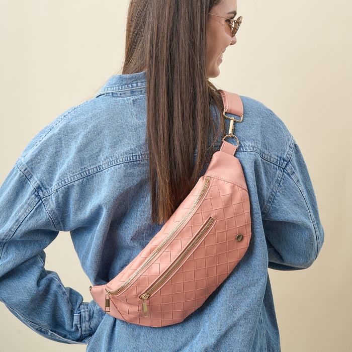 Woman with long brown hair wears sunglasses, a blue denim jacket, and The Darling Effect's Rose Trendy Luxe Belt Bag with gold zippers, posing with her back to the camera against a beige background.
