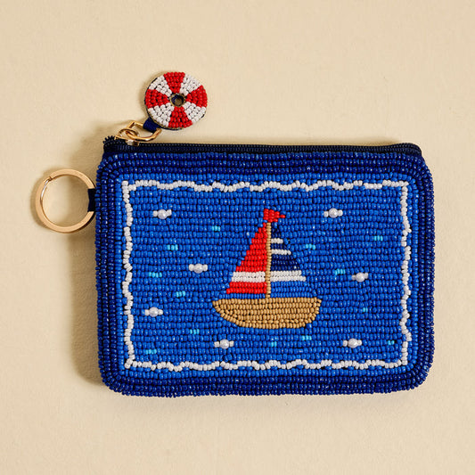 The Darling Effect’s Sail Away Handcrafted Beaded Pouch features a blue sailboat design, red and white life preserver charm, and gold keyring—a stylish artisan accessory for nautical flair.