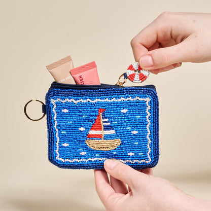 The Darling Effect's Sail Away Handcrafted Beaded Pouch in blue, held by two hands, features a sailboat design and comes with a small life preserver keychain attached to the zipper.