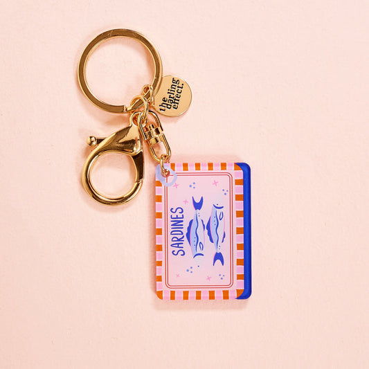 The Sardines Acrylic Keychain by The Darling Effect features a sardine can design with two blue sardines on a pink background, orange stripes, a gold keyring, and a logo charm—perfect for acrylic keychain fans.