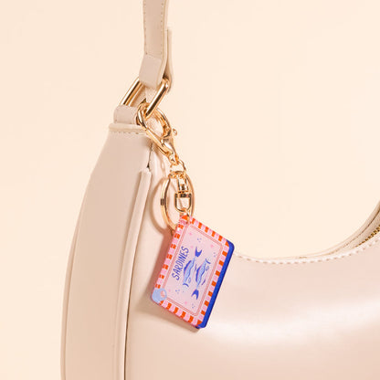 Close-up of The Darling Effect’s Sardines Acrylic Keychain, featuring pink and red stripes and fish illustrations, attached to a cream handbag with a gold clasp. The beige background enhances this stylish accessory's charm.