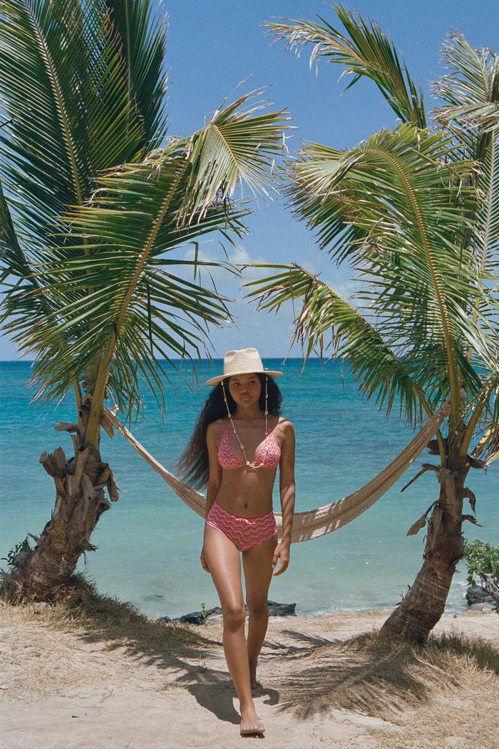 Model wearing Seashells Fedora hat, walking on beach with palm trees in background