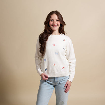 A young woman with long brown hair smiles against a beige background, wearing The Darling Effect’s Seaside Haven Refined Knit Sweater—an elegant piece featuring small, colorful embroidered designs—paired with light blue jeans.