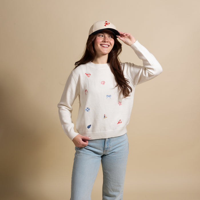 A smiling woman wears The Darling Effect’s Seaside Haven Refined Knit Sweater—an elegant piece with colorful embroidery—paired with light blue jeans and a beige cap, standing against a plain beige background.