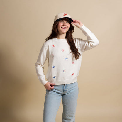 A smiling woman wears The Darling Effect’s Seaside Haven Refined Knit Sweater—an elegant piece with colorful embroidery—paired with light blue jeans and a beige cap, standing against a plain beige background.