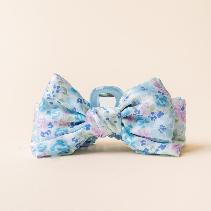 The-darling-effect’s Secret Garden Blue Satin Bow Claw Clip features a pastel blue and pink floral design, ideal for updos, and is showcased against a plain beige background.