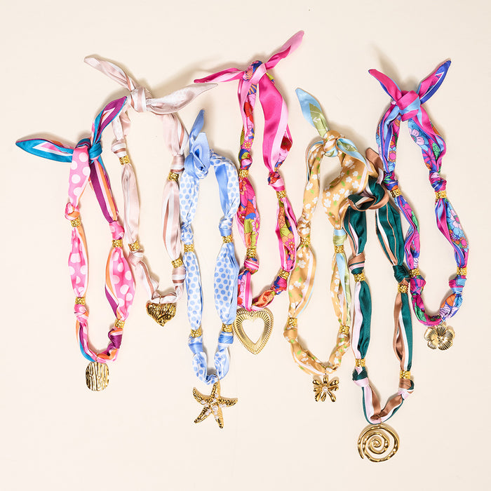 Six colorful scarf necklaces with artful patterns and various gold charms—heart, starfish, flower, shell, swirl, and disc—are laid side by side on a light background.