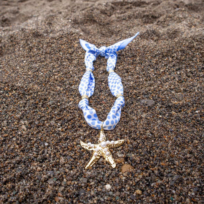 The Darling Effect Shore Thing Make a Statement Scarf Necklace, featuring blue and white polka dots and a gold starfish charm, is displayed on brown sand.