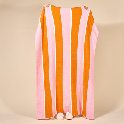 A person holds up The Darling Effect's Sicily Stripes Pink Cozy Dreams Luxe Blanket, featuring wide pink, orange, and white stripes. Only their hands and feet in white slippers are visible against a beige background.