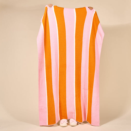 A person holds up The Darling Effect's Sicily Stripes Pink Cozy Dreams Luxe Blanket, featuring wide pink, orange, and white stripes. Only their hands and feet in white slippers are visible against a beige background.