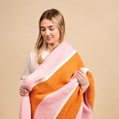 A young woman drapes The Darling Effect's Sicily Stripes Pink Cozy Dreams Luxe Blanket over her shoulders, showcasing its pink, orange, and white stripes. She stands against a beige background, smiling gently in a white top.