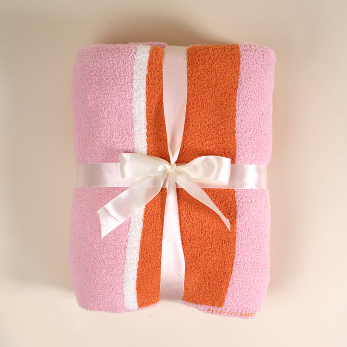 The Sicily Stripes Pink Cozy Dreams Luxe Blanket by The Darling Effect features pink, orange, and white stripes, a plush microfiber texture, and comes folded with a white satin bow, displayed on a light beige background.