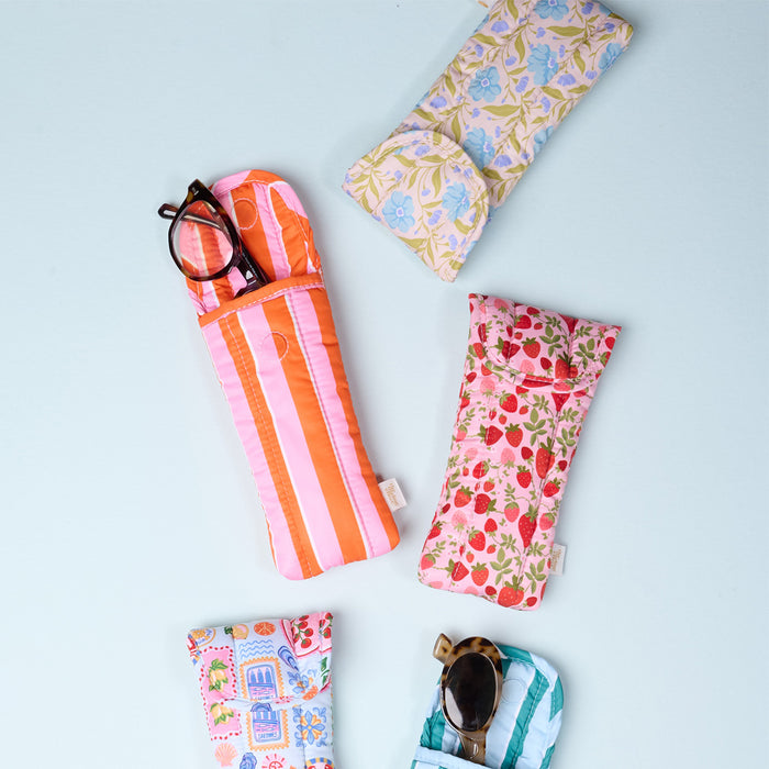Five eyeglass cases, including The Darling Effect Sicily Stripes Pink Puffy Sunglasses Case, are arranged on a light blue surface. Two pairs of glasses peek out from floral, striped, and pastel fruit-themed designs.