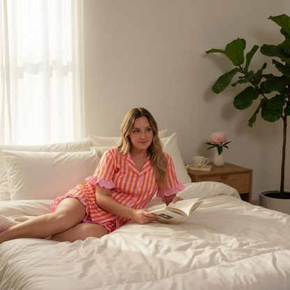 A woman in The Darling Effect’s Sicily Stripes Pink Slumber Party Pajama Shorts Set relaxes on a bed with white bedding, holding an open book. A nightstand with a cup, saucer, flower, and potted plant sits nearby as sunlight filters through the window.