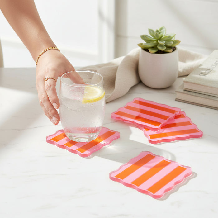 A hand sets a glass of water with lemon on The Darling Effect Sicily Stripes Pink Table Talk Acrylic Coaster, while three more coasters sit nearby beside a potted succulent and stacked books, adding playful décor to the table.