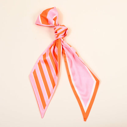 The Darling Effect’s Sicily Stripes Pink Twirl & Tie Scarf Accessory, featuring pink and orange diagonal and chevron stripes, is shown tied in a loose knot on a light beige background.