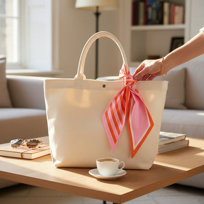 A cream tote featuring The Darling Effect's Sicily Stripes Pink Twirl & Tie Scarf Accessory is placed on a coffee table next to coffee, sunglasses, and books in a sunlit living room. A hand reaches for this stylish piece.