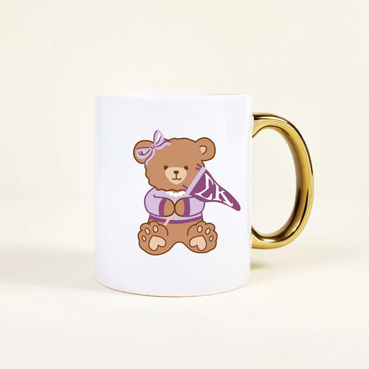 The Sigma Kappa 11 oz Cozy Cute Ceramic Mug by The Darling Effect features a brown teddy bear in a purple bow and sweater, holding an “LK” pennant. The white mug has a gold handle for stylish flair.