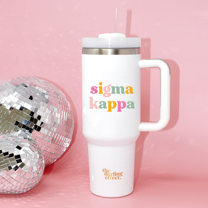 The Sigma Kappa 40 oz Take Me Everywhere White Tumbler by The Darling Effect, featuring “sigma kappa” in colorful letters and the brand’s logo, sits beside a silver disco ball on a pink background—making this insulated drinkware a fun and functional companion for all your adventures.