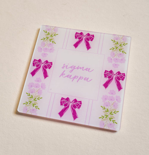 The Darling Effect’s Sigma Kappa Acrylic Coaster features pink bows and purple flowers on a light background with “Sigma Kappa” in cursive—an ideal sorority gift or stylish addition to your coaster collection.