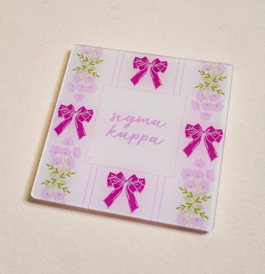 The Darling Effect’s Sigma Kappa Acrylic Coaster features pink bows and purple flowers on a light background with “Sigma Kappa” in cursive—an ideal sorority gift or stylish addition to your coaster collection.