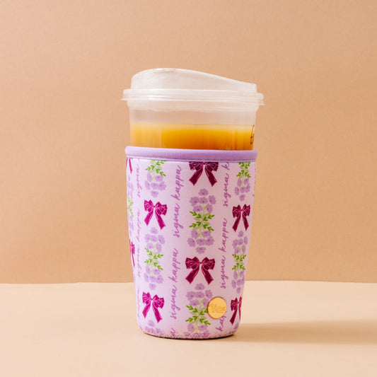 The Sigma Kappa Large Sip Savvy Drink Sleeve by The Darling Effect wraps a plastic cup of orange drink in light purple neoprene with purple bows, green leaves, and "kappa kappa" script—perfect for sorority pride. The cup rests on a beige background.