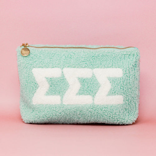 The Darling Effect’s Sigma Sigma Sigma Aqua Teddy Letter Pouch features plush mint sherpa, a gold zipper, round tag, and three white ΣΣΣ letters—ideal as a chic carry-on accessory. Styled with a light pink background.