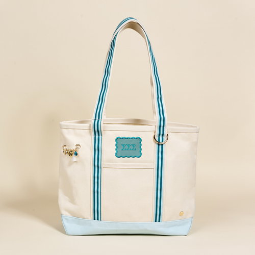 The Sigma Sigma Sigma Canvas Tote Bag by The Darling Effect is a cream-colored, durable canvas tote with light blue accents, striped handles, a blue Greek letters patch, and a decorative charm on the handle. Background: neutral beige.