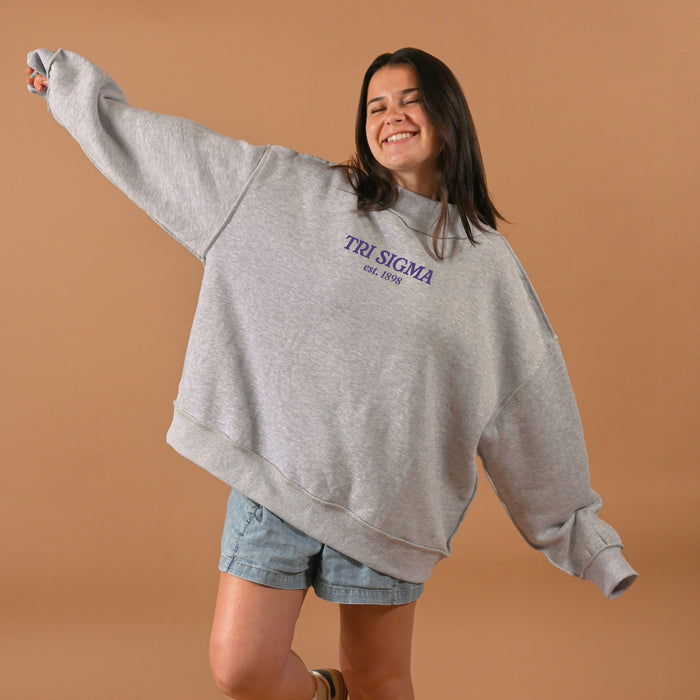 A smiling woman wears The Darling Effect’s Sigma Sigma Sigma Chapter Mockneck Sweatshirt in L/XL gray with denim shorts, arms outstretched against a plain tan background.