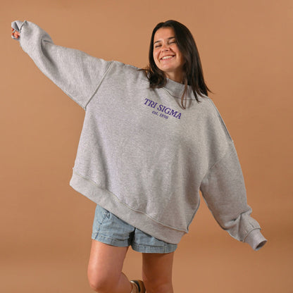 A smiling woman wears The Darling Effect’s Sigma Sigma Sigma Chapter Mockneck Sweatshirt in L/XL gray with denim shorts, arms outstretched against a plain tan background.