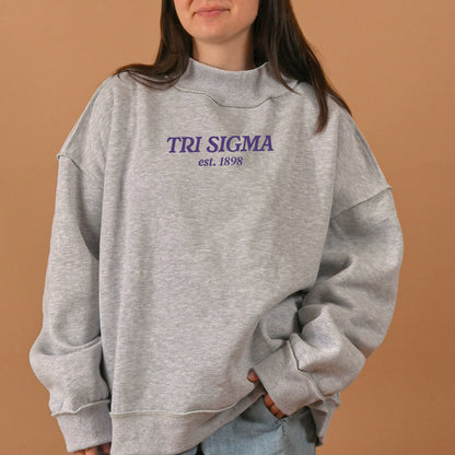 A person wearing an oversized, light grey Sigma Sigma Sigma Chapter Mockneck Sweatshirt L/XL by The Darling Effect, featuring "TRI SIGMA est. 1898" printed in purple text on the front, stands against a tan background.
