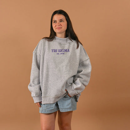 A woman with long brown hair and a relaxed smile stands before a tan background, wearing The Darling Effect's Sigma Sigma Sigma Chapter Mockneck Sweatshirt S/M in purple text and denim shorts.