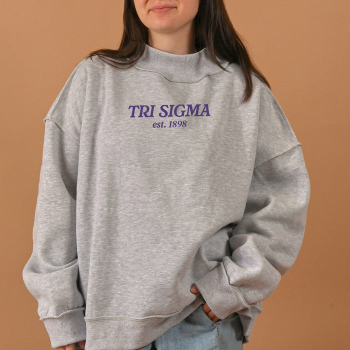 A woman stands against a plain tan background wearing The Darling Effect Sigma Sigma Sigma Chapter Mockneck Sweatshirt (size S/M), an oversized gray sweatshirt with "TRI SIGMA est. 1898" in purple and blue lettering on the front and a cozy mock neck design.