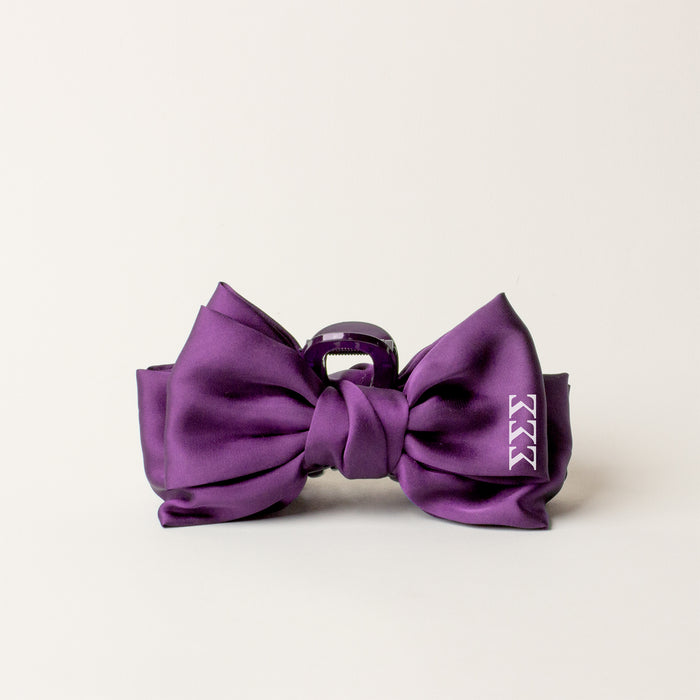 The Darling Effect’s Sigma Sigma Sigma Satin Bow Claw Clip in satin purple features a shiny finish and a small white triple "K" logo—an elegant hair accessory perfect for updos, shown against a plain light background.