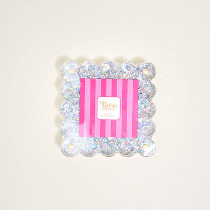 The Silver Glitter Little Moments Acrylic Picture Frame by The Darling Effect features a square, scalloped edge with glitter, a 2.5x2.5 photo space, pink and white striped insert, "the darling effect" text, and a magnetic closure.