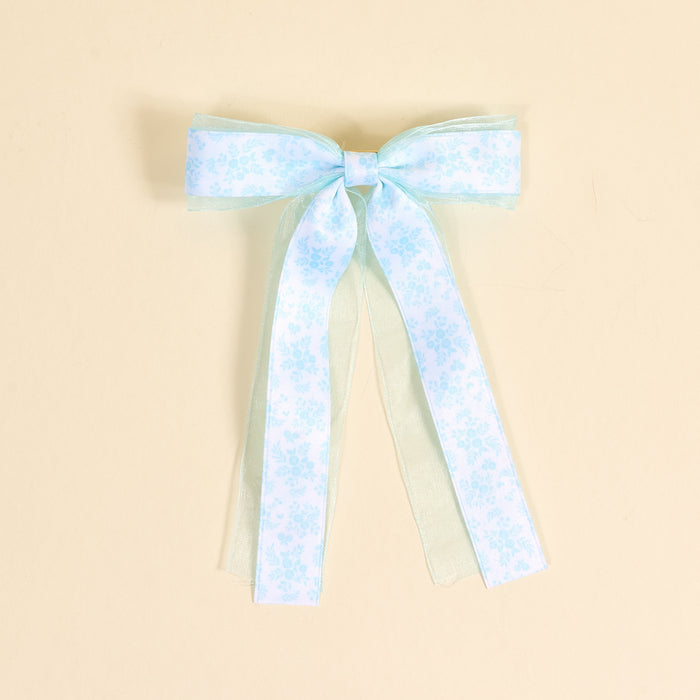 The Darling Effect’s Something Blue Lovely Layered Hair Bow features subtle floral patterns and sheer ribbon tails, shown on a beige background—an elegant addition to any hair accessory collection.
