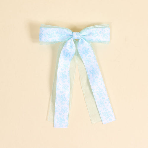 The Darling Effect’s Something Blue Lovely Layered Hair Bow features subtle floral patterns and sheer ribbon tails, shown on a beige background—an elegant addition to any hair accessory collection.