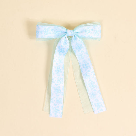 The Darling Effect’s Something Blue Lovely Layered Hair Bow features subtle floral patterns and sheer ribbon tails, shown on a beige background—an elegant addition to any hair accessory collection.