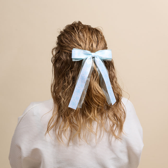 A person with wavy, shoulder-length light brown hair wears The Darling Effect's Something Blue Lovely Layered Hair Bow, featuring long tails, paired with a white top against a beige background.