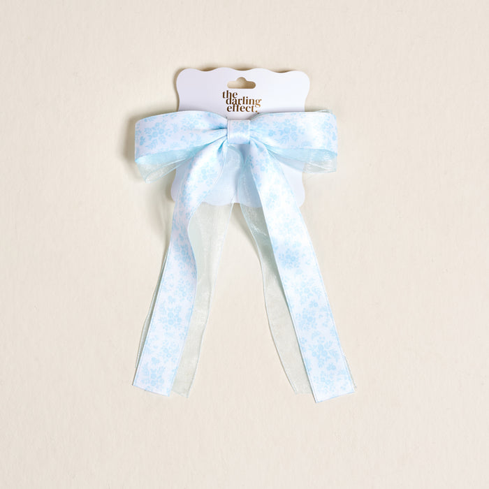 The Darling Effect's Something Blue Lovely Layered Hair Bow features a light blue hue with a white floral pattern and sheer ribbon tails, adding charm to your accessories. Shown on a cream background.