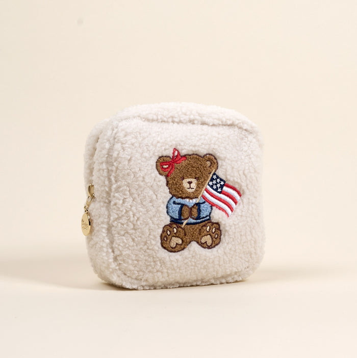 The Darling Effect’s Square Star Spangled Bear Teddy Pouch is a small, cream sherpa zip pouch with a gold tag and features an embroidered bear with an American flag—a cute addition to your teddy pouches collection.