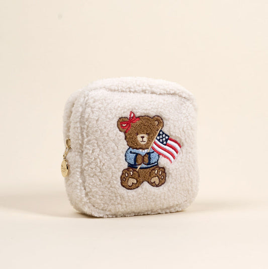 The Darling Effect’s Square Star Spangled Bear Teddy Pouch is a small, cream sherpa zip pouch with a gold tag and features an embroidered bear with an American flag—a cute addition to your teddy pouches collection.