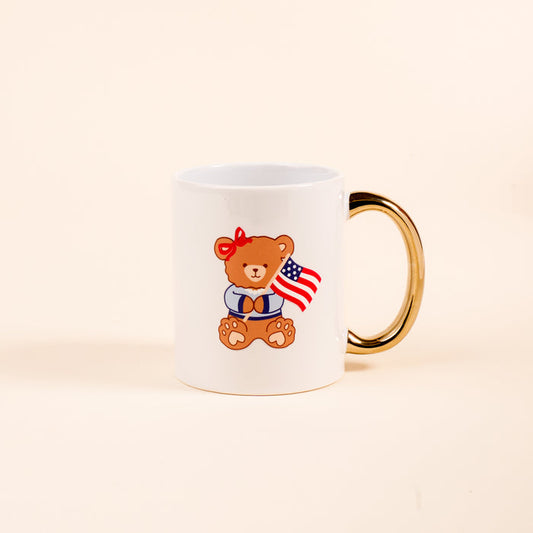 Sip in style with The Darling Effect’s Star Spangled Bear 11 oz Cozy Cute Ceramic Mug, featuring a white ceramic design, golden handle, and an adorable teddy bear with a red bow and American flag on a bone-shaped mat.