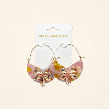 The Darling Effect's Sunset Kiss Marble Bloom Hoop Earrings are lightweight, half-moon shaped with a pink and yellow marbled design and feature a pink crystal bow on the front, displayed on a branded card.