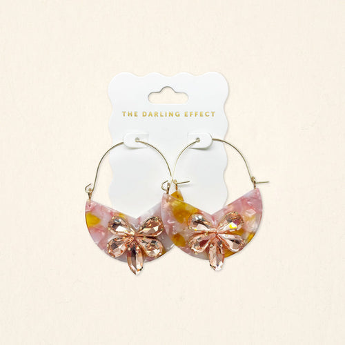 The Darling Effect's Sunset Kiss Marble Bloom Hoop Earrings are lightweight, half-moon shaped with a pink and yellow marbled design and feature a pink crystal bow on the front, displayed on a branded card.