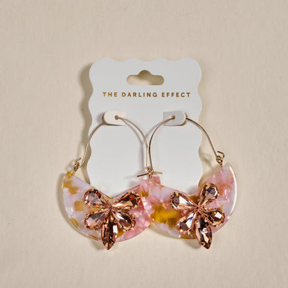The Darling Effect Sunset Kiss Marble Bloom Hoop Earrings feature crescent-shaped pink, gold, and white acrylic with large pink rhinestone flower accents. Lightweight statement earrings displayed on a branded card, perfect for adding flair to any outfit.
