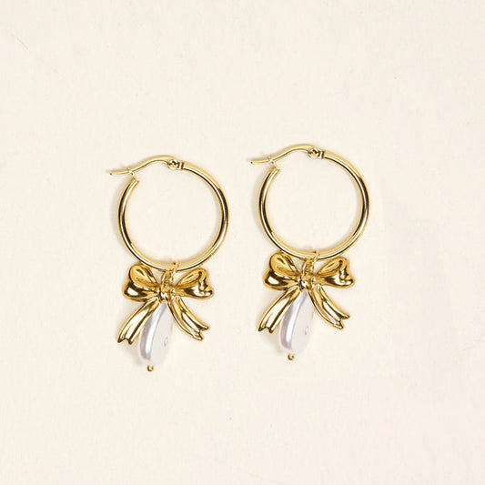 The Darling Effect’s Sweetheart Pearl Signature Hoop Earrings: lightweight gold hoops adorned with delicate bow charms and white teardrop-shaped beads, shown on a light beige background.
