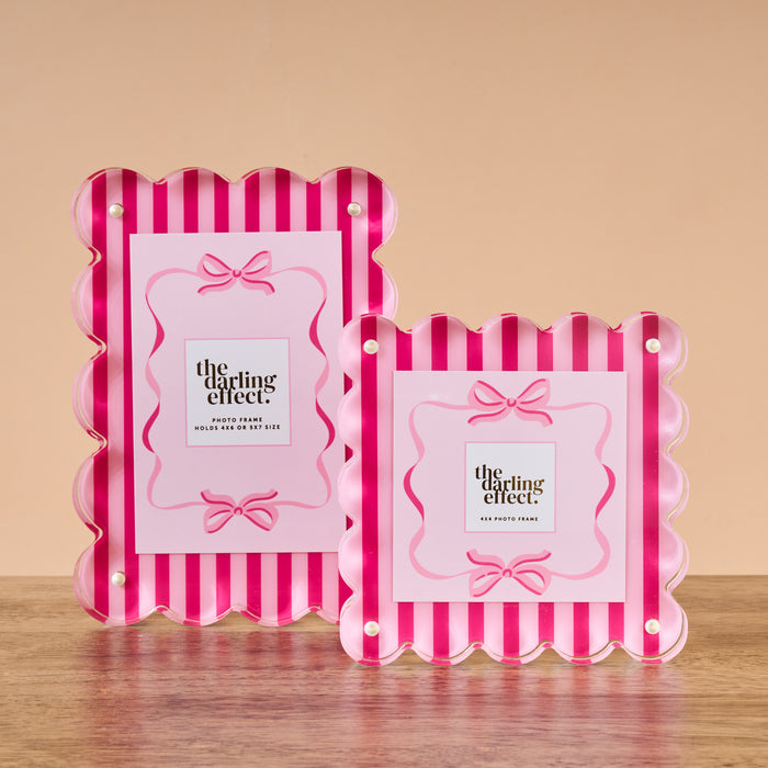Two Sweetheart Stripes Rectangle Scalloped Acrylic Frames by The Darling Effect, featuring pink and red stripes with bow designs, are displayed on a wooden surface against a plain beige background.