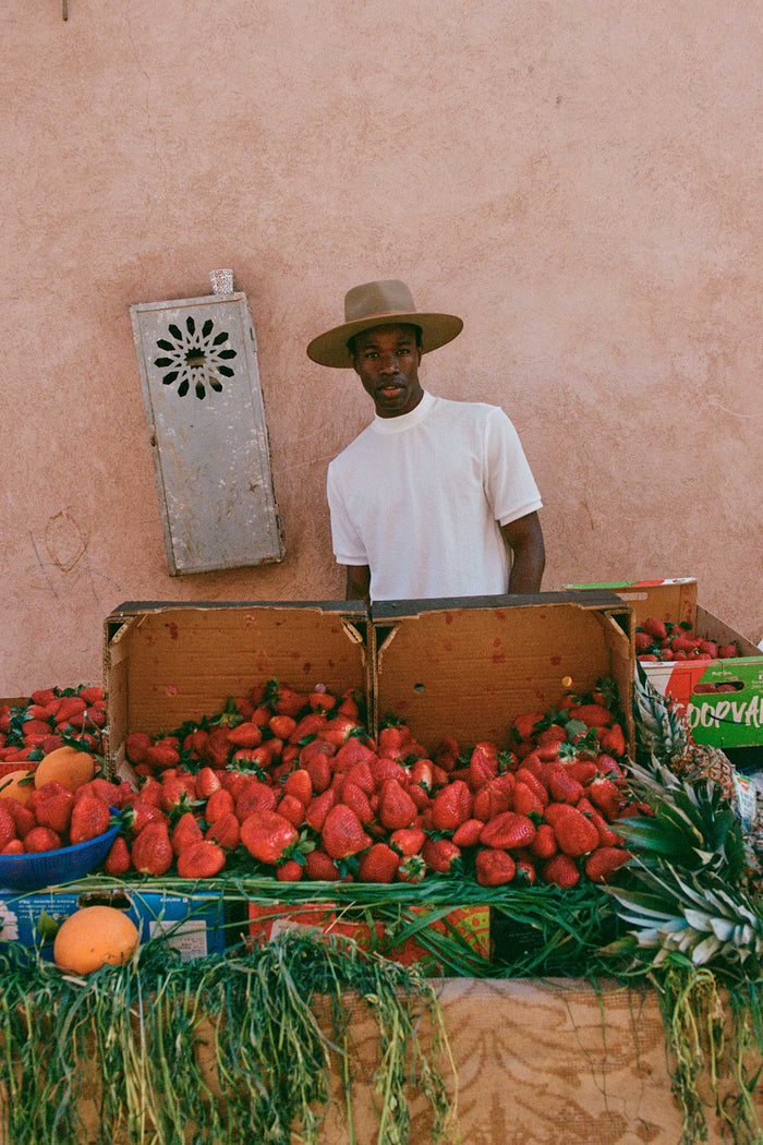 A man wearing a brown wool fedora hat at a fruit market, surrounded by strawberries and other fruits.